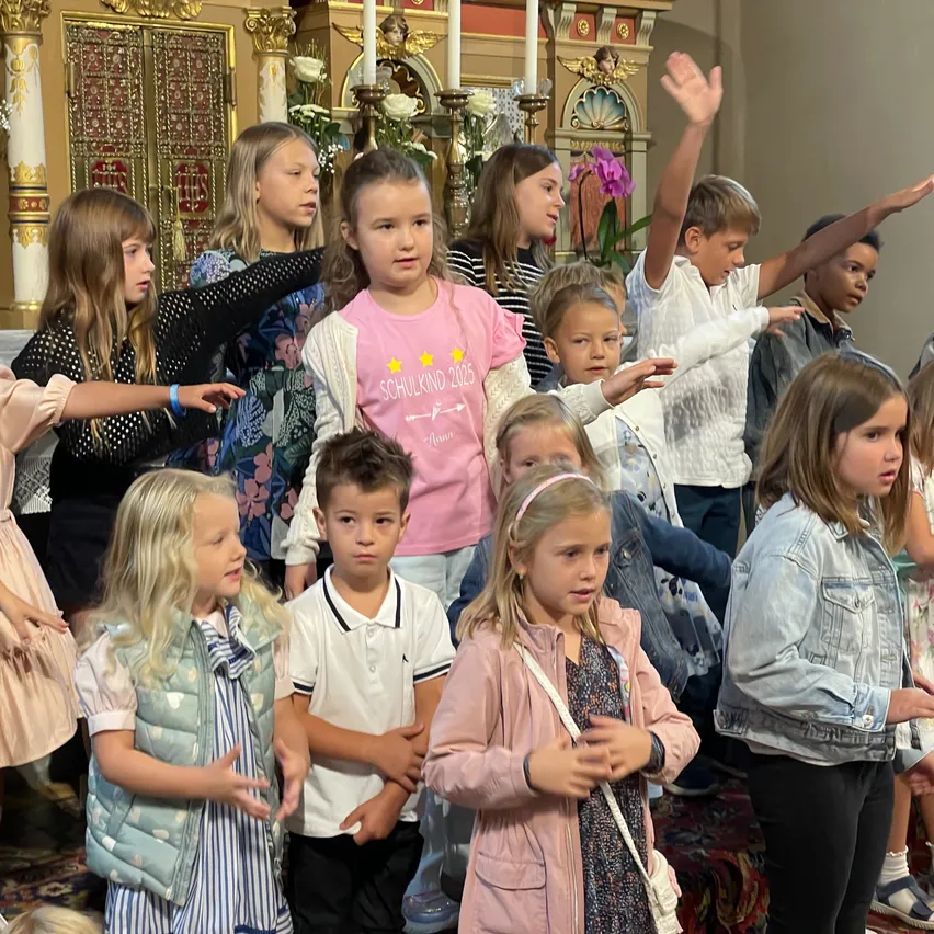 Eine Gruppe von Kindern in verschiedenen Outfits steht in einer Kirche, einige mit erhobenen Armen, wahrscheinlich bei einer religiösen Veranstaltung oder einem Gottesdienst.