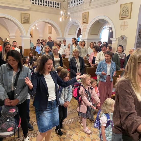 Eine Gruppe von Menschen ist in einer Kirche versammelt, möglicherweise für einen Gottesdienst oder eine Veranstaltung. Sie sind lässig gekleidet und scheinen in Gespräche vertieft zu sein. Einige halten Kinder. Die Kirche hat gewölbte Eingänge und eine Kronleuchter.