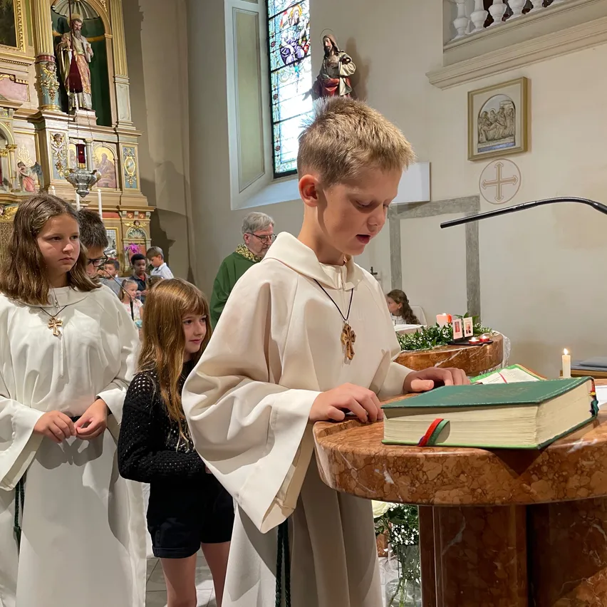 Ein junger Junge steht an einem Podest und liest aus einem großen grünen Buch in einer Kirche. Ein Mädchen in einem schwarzen Kleid und ein anderes in einem weißen Gewand stehen in der Nähe. Im Hintergrund beobachtet ein Mann mit Brille.