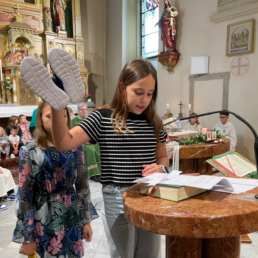 Ein junges Mädchen hält ihre Schuhe hoch, während ein anderes Mädchen in einer Kirche zuschaut. Papiere und Bücher sind auf dem Tisch.