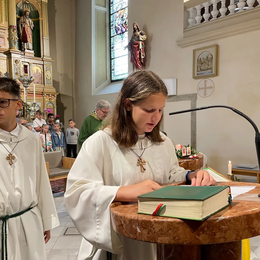 Ein junges Mädchen in einer weißen Robe liest aus einem Buch auf einem Podium in einer Kirche, während eine andere Person in einer weißen Robe hinter ihr steht. Dahinter sind eine Statue und ein Fenster zu sehen.