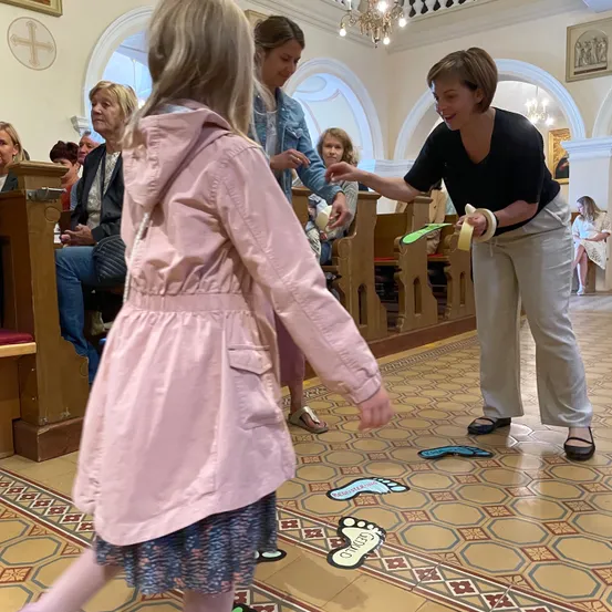 Ein junges Mädchen in einem rosa Mantel spielt ein Spiel auf dem Kirchenboden, während eine Frau in Schwarz ihre Hand hält. Mehrere Personen sitzen auf hölzernen Bänken hinter ihnen.