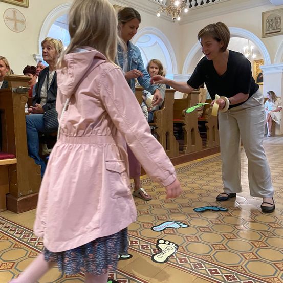 Ein junges Mädchen in einem rosa Mantel spielt ein Spiel auf dem Kirchenboden, während eine Frau in Schwarz ihre Hand hält. Mehrere Personen sitzen auf hölzernen Bänken hinter ihnen.