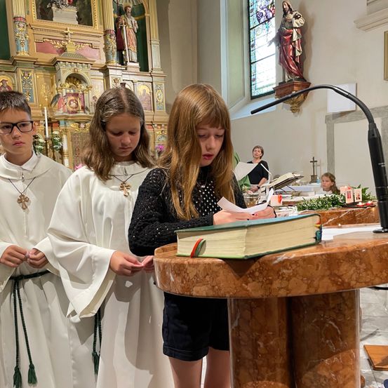 Drei Kinder, zwei in weißen Roben und eines in einem schwarzen Shirt, stehen vor einem Pult. Sie sehen ein Buch an. Vor dem Buch steht ein Mikrofon.