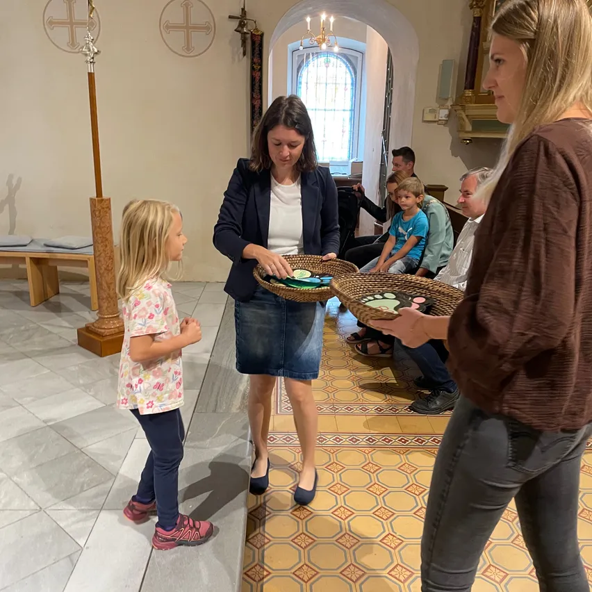 In einer Kirche gibt eine Frau, die einen Korb hält, diesen an ein Kind weiter, das neben einer anderen Frau steht, während mehrere Personen auf Bänken im Hintergrund sitzen.