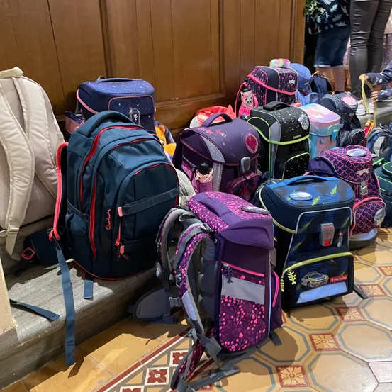 Viele bunte Rucksäcke und Taschen sind auf dem gefliesten Boden gestapelt, mit einer hölzernen Wand im Hintergrund.