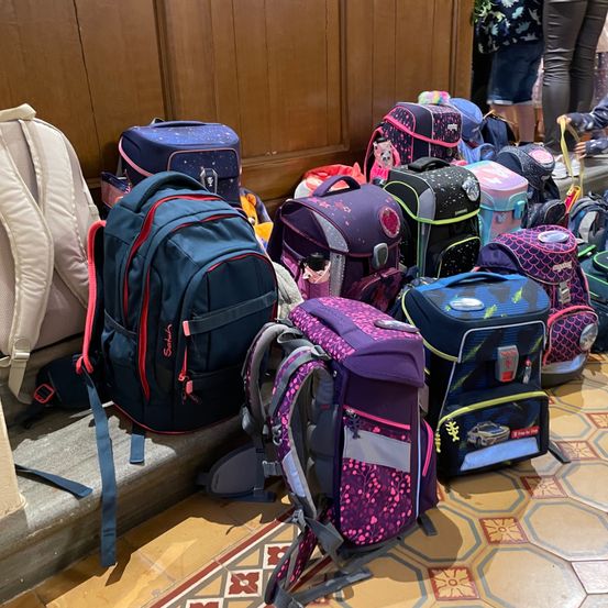 Viele bunte Rucksäcke und Taschen sind auf dem gefliesten Boden gestapelt, mit einer hölzernen Wand im Hintergrund.