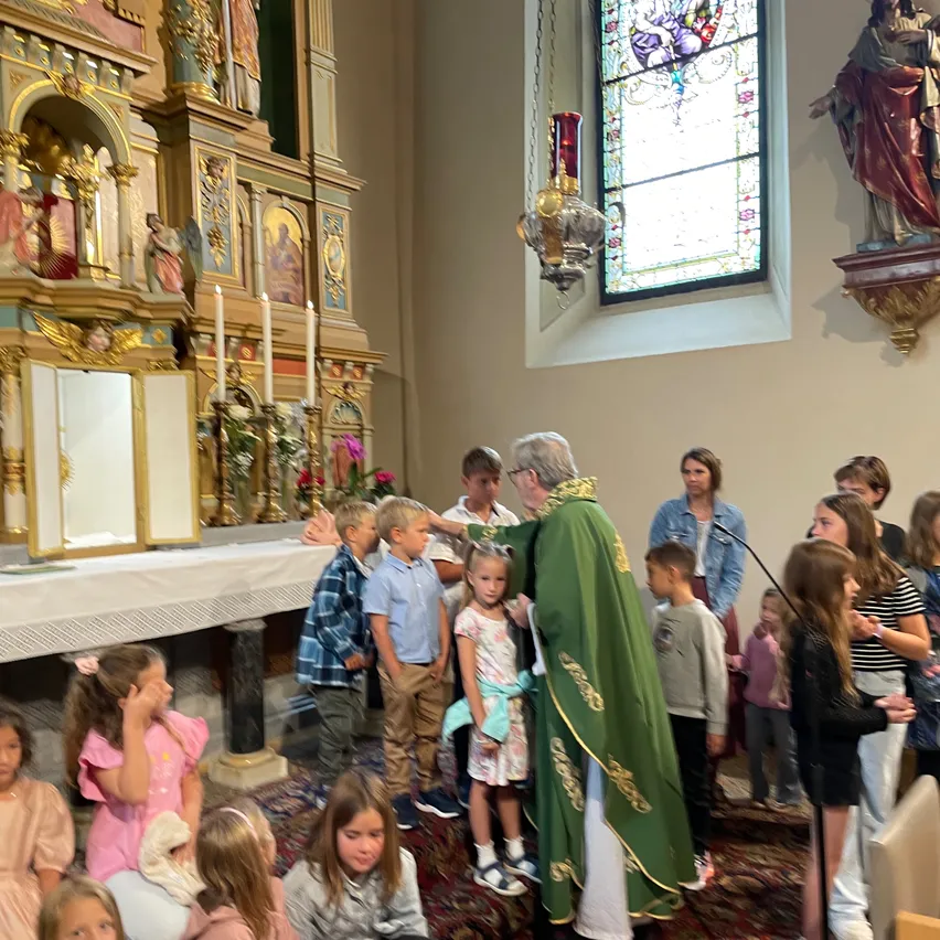 Ein Priester in einem grünen Gewand steht in einer Kirche und führt Kinder, die ihm aufmerksam zuschauen. Hinter dem Priester steht ein Altar mit Kerzen und seitlich befindet sich ein Buntglasfenster.