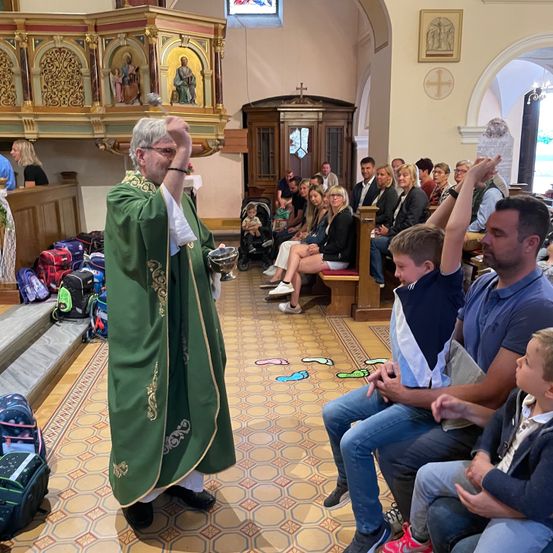 Ein Priester steht in einer Kirche, hält einen Kelch und ist von einer Gemeinde mit Kindern umgeben, während ein Kinderwagen in der Nähe steht und Taschen auf dem Boden liegen.