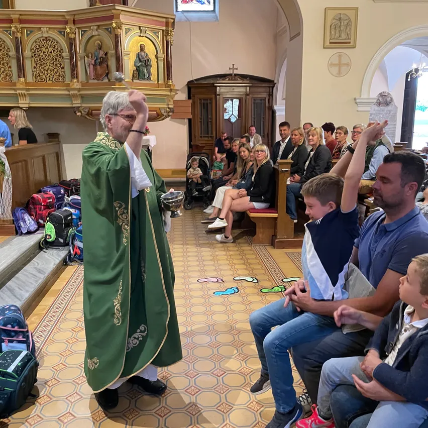 Ein Priester steht in einer Kirche, hält einen Kelch und ist von einer Gemeinde mit Kindern umgeben, während ein Kinderwagen in der Nähe steht und Taschen auf dem Boden liegen.
