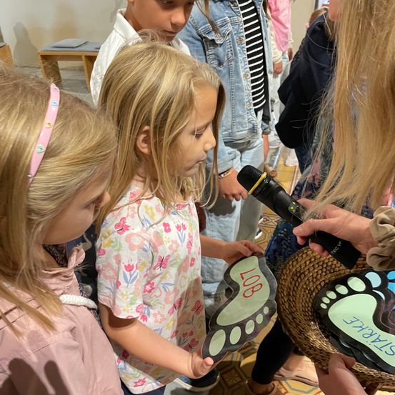 Eine Gruppe von Kindern, darunter eine mit einem Mikrofon, versammelt sich um eine Person, die einen Korb hält. Das Mädchen in der Mitte hält eine fußförmige Aussparung mit der Aufschrift '108'. Sie scheinen an einer Veranstaltung teilzunehmen.