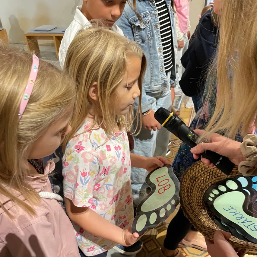 Eine Gruppe von Kindern, darunter eine mit einem Mikrofon, versammelt sich um eine Person, die einen Korb hält. Das Mädchen in der Mitte hält eine fußförmige Aussparung mit der Aufschrift '108'. Sie scheinen an einer Veranstaltung teilzunehmen.