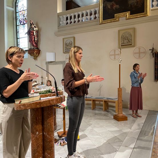 Zwei Frauen sprechen an einem Pult in einer Kirche mit einer Statue und Kreuzen. Eine dritte Frau steht hinter ihnen und klatscht.