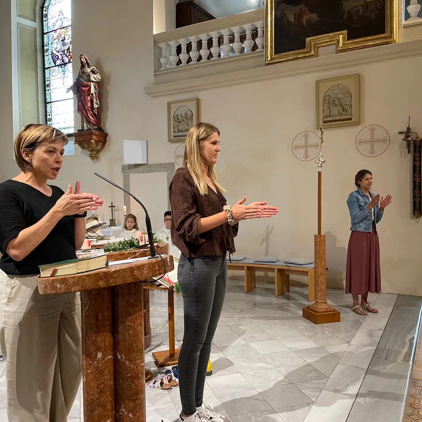 Zwei Frauen sprechen an einem Pult in einer Kirche mit einer Statue und Kreuzen. Eine dritte Frau steht hinter ihnen und klatscht.