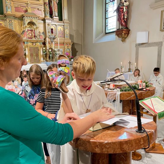 Eine Frau hält die Sandalen eines Kindes, während er von einem Buch am Altar einer Kirche liest, umgeben von Kindern und Kerzen.