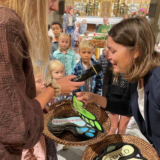 Eine Gruppe von Kindern und Erwachsenen versammelt sich in einer Kirche. Zwei Frauen halten Körbe mit Schmetterlingsdesigns. Eine Frau hält ein Mikrofon und scheint zu sprechen.