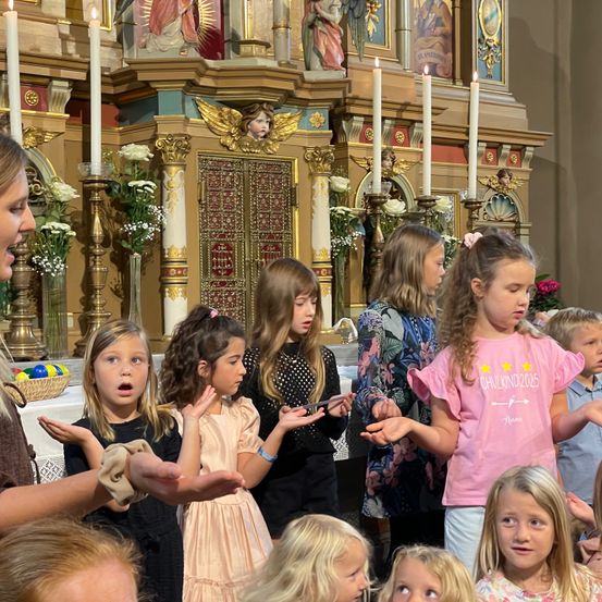 Eine Gruppe von Kindern, darunter Mädchen in Kleidern und Jungen in Hemden, singen vor einem Kirchenaltar, Kerzen und Blumen.