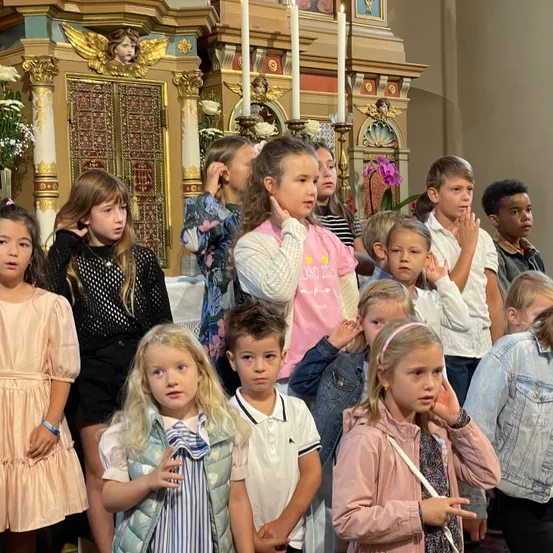 Eine Gruppe von Kindern steht in einer Kirche und hört aufmerksam der Predigt zu, wobei einige ihre Ohren bedecken. Eine goldene Engelsstatue schmückt den Altar.
