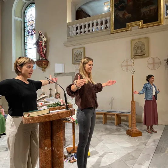 Drei Frauen stehen in einer Kirche, eine spricht in ein Mikrofon am Altar. Eine weitere Frau steht in der Nähe mit ausgestreckten Armen. Eine dritte Frau steht rechts. Hinter ihnen schmücken Bänke, eine Statue und gerahmte Bilder die Wände.