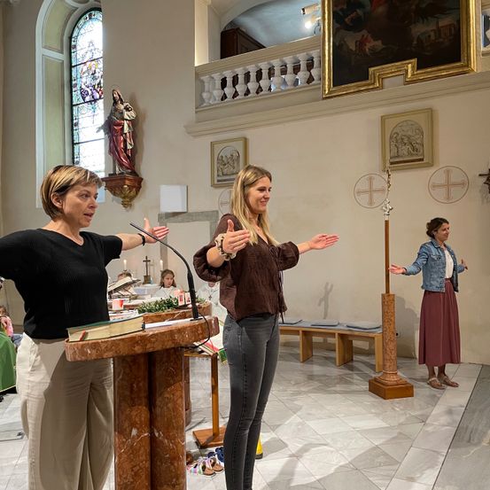 Drei Frauen stehen in einer Kirche, eine spricht in ein Mikrofon am Altar. Eine weitere Frau steht in der Nähe mit ausgestreckten Armen. Eine dritte Frau steht rechts. Hinter ihnen schmücken Bänke, eine Statue und gerahmte Bilder die Wände.