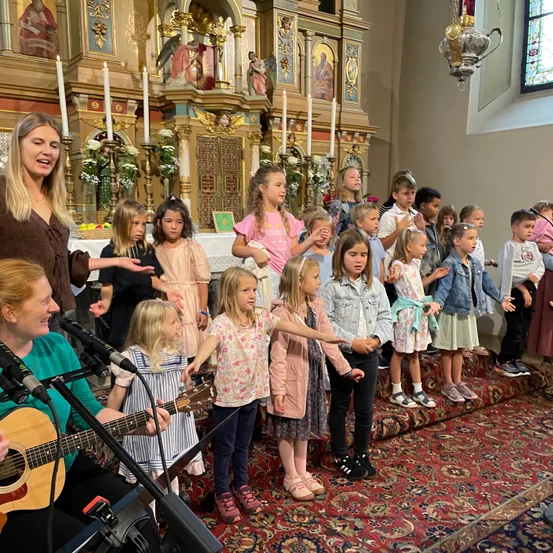 Eine Gruppe von Kindern und zwei Frauen steht vor einem Altar in einer Kirche, singt, während eine Frau Gitarre spielt und die andere spricht.