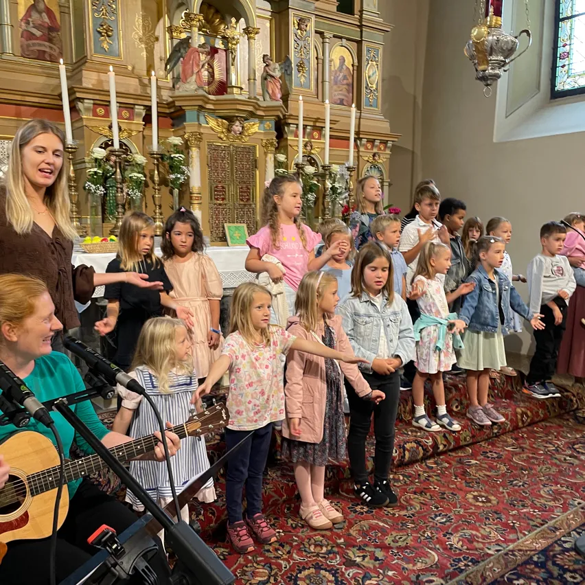 Eine Gruppe von Kindern und zwei Frauen steht vor einem Altar in einer Kirche, singt, während eine Frau Gitarre spielt und die andere spricht.