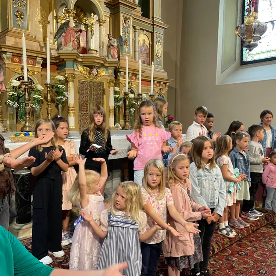 Eine Gruppe junger Kinder und einige Erwachsene stehen in einer Kirche und blicken auf den Altar mit brennenden Kerzen. Die Kinder klatschen.