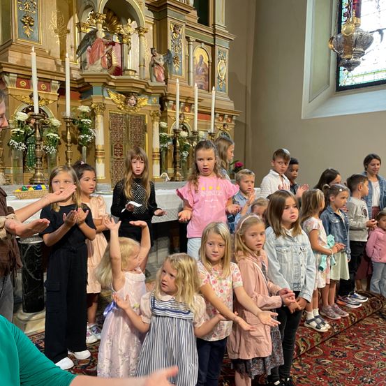 Eine Gruppe junger Kinder und einige Erwachsene stehen in einer Kirche und blicken auf den Altar mit brennenden Kerzen. Die Kinder klatschen.