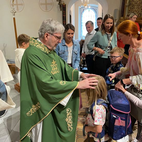 Ein Priester segnet ein Kind in einer Kirche, während mehrere Leute zusehen. Der Priester trägt einen grünen Umhang.