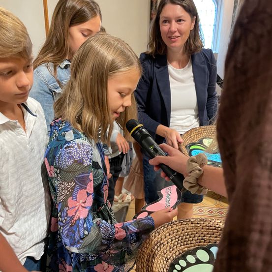 Ein junges Mädchen spricht in ein Mikrofon, das eine Frau hält. Hinter ihr schauen ein Junge und ein Mädchen zu, während eine andere Frau das Mädchen anlächelt.