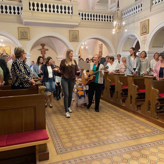 Mehrere Menschen versammeln sich in einer Kirche, einige spielen Gitarre, mit Kirchenbänken und einem Altar im Hintergrund.
