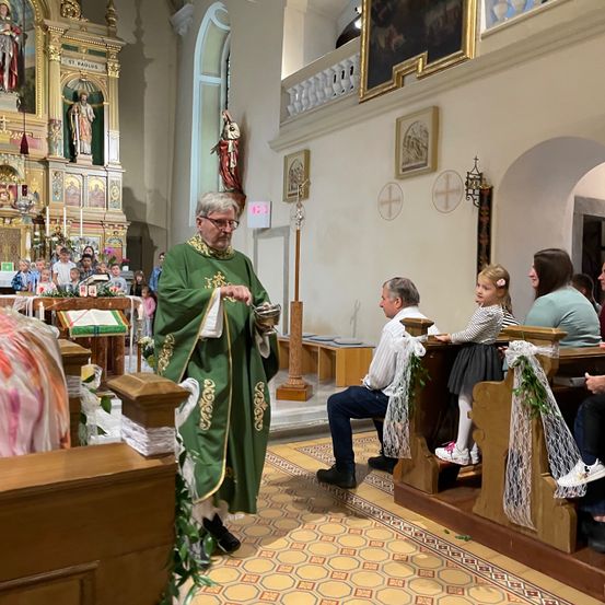 Ein älterer Priester in einer grünen Robe steht in einer Kirche, wo mehrere Menschen auf Bänken sitzen. Vor ihm sitzen Kinder auf dem Altar mit einer Statue und Kerzen.