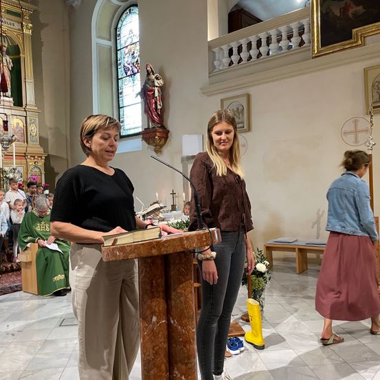 Zwei Frauen stehen vor einem Podest in einer Kirche und lesen aus einem Buch. Ein Mikrofon steht vor ihnen. Dahinter sitzen eine Gruppe von Menschen.