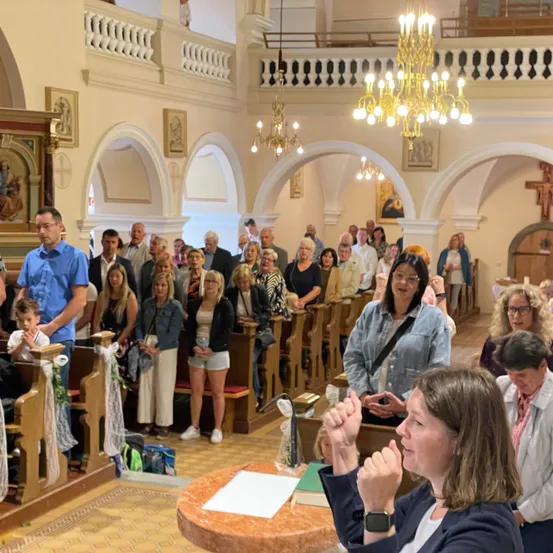 Ein Gottesdienst mit einem Publikum aus Männern und Frauen, die auf Kirchenbänken sitzen. Einige klatschen, während eine Frau vor einem Tisch steht.