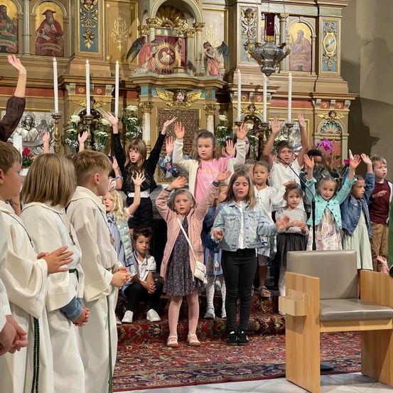Eine Gruppe von Kindern in einer Kirche, die ihre Hände heben, einige stehend und andere sitzend, vor einem Altar mit Kerzen und religiösen Dekorationen.