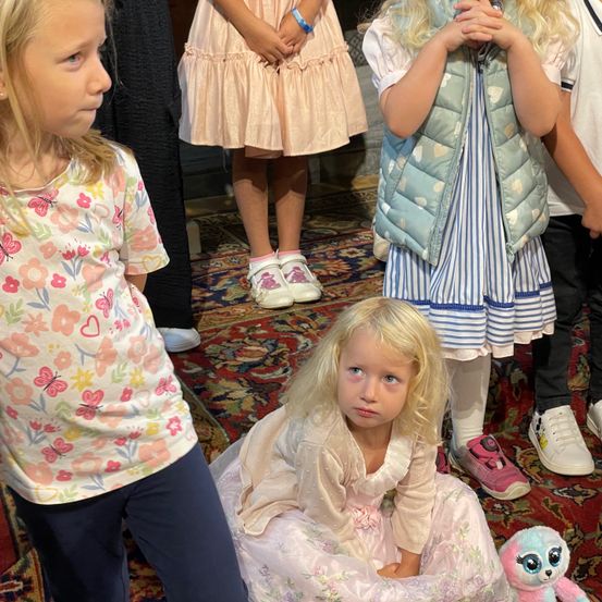 Mehrere Kinder stehen zusammen auf einem Teppich. Ein Mädchen vorne hat blonde Haare und trägt ein Blumenhemd. Ein Mädchen neben ihr trägt ein rosa Kleid. Ein weiteres Mädchen hinter ihr trägt einen rosa Schuh.