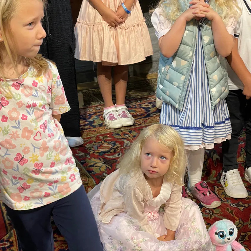Mehrere Kinder stehen zusammen auf einem Teppich. Ein Mädchen vorne hat blonde Haare und trägt ein Blumenhemd. Ein Mädchen neben ihr trägt ein rosa Kleid. Ein weiteres Mädchen hinter ihr trägt einen rosa Schuh.