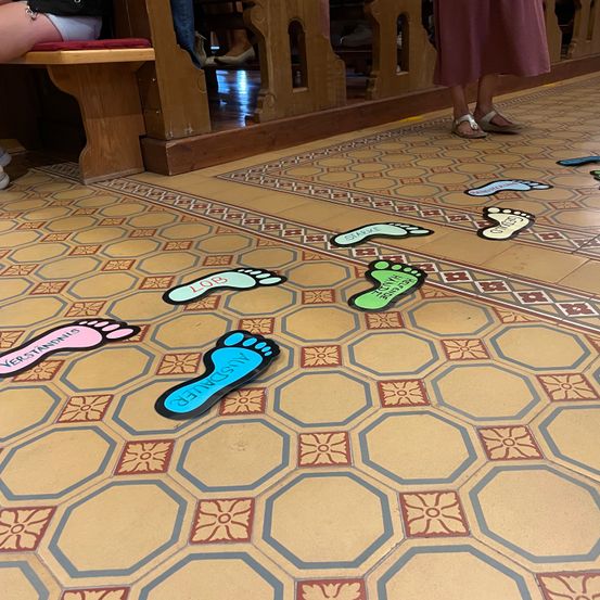 Sechseckige Fliesen bedecken den Boden, verziert mit bunten Fußabdrücken, die zu einer Bank führen. Menschen sitzen auf der Bank, einer steht in der Nähe.