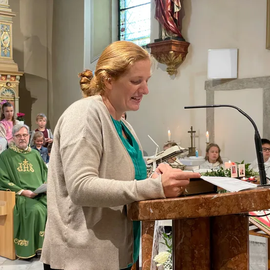 Eine Frau liest aus einem Buch an einer Kanzel in einer Kirche mit Kerzen und einer Statue im Hintergrund.