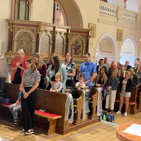 Eine Gruppe von Menschen steht in einer Kirche, einige beten. Sie stehen vor dem Altar, mit Bänken und einem Gemälde von Jesus. Die Atmosphäre ist feierlich.