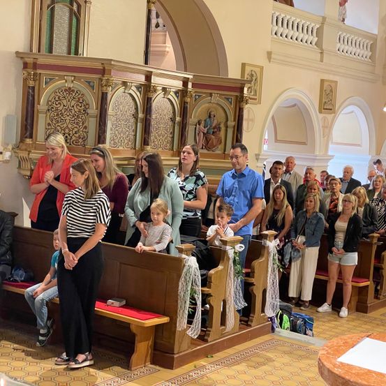 Eine Gruppe von Menschen steht in einer Kirche, einige beten. Sie stehen vor dem Altar, mit Bänken und einem Gemälde von Jesus. Die Atmosphäre ist feierlich.