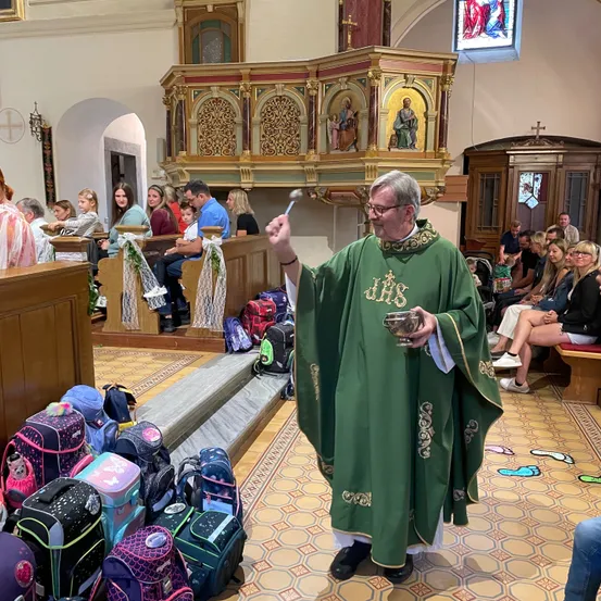 Ein Priester steht vor einem Kirchenaltar, hält einen Kelch und vor ihm liegen Rucksäcke. Im Hintergrund sitzen Menschen, einige mit Brille.