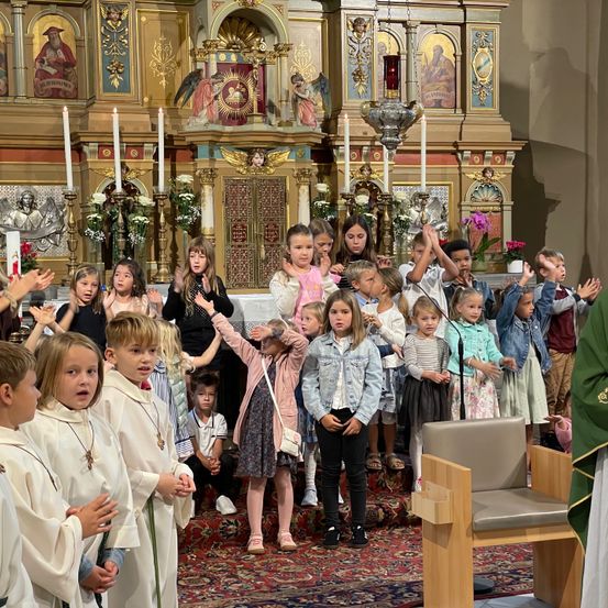 Eine Gruppe von Kindern in weißen Gewändern steht in einer Kirche, vor einem Altar mit Kerzen und einem Priester. Einige halten die Hände erhoben, andere schauen umher, während ein Stuhl in der Nähe steht.