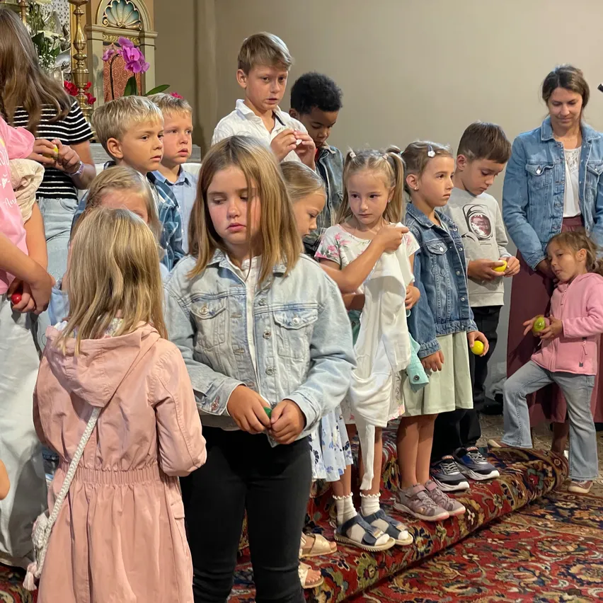 Eine Gruppe von Kindern und Erwachsenen, darunter junge Mädchen und Jungen, stehen zusammen, einige halten Äpfel. Sie scheinen an einem Gottesdienst teilzunehmen, wobei einige beten und andere nach unten schauenn.