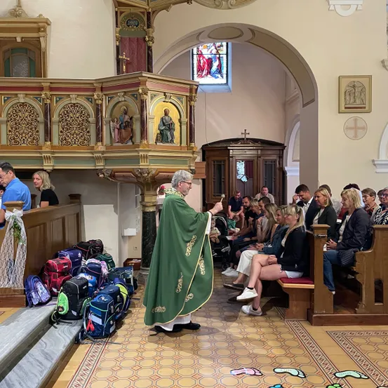 Ein Priester steht vor einer Gemeinde in einer Kirche und hält eine Rede. Taschen und Rucksäcke liegen auf dem Boden, und die Menschen sitzen auf Bänken.