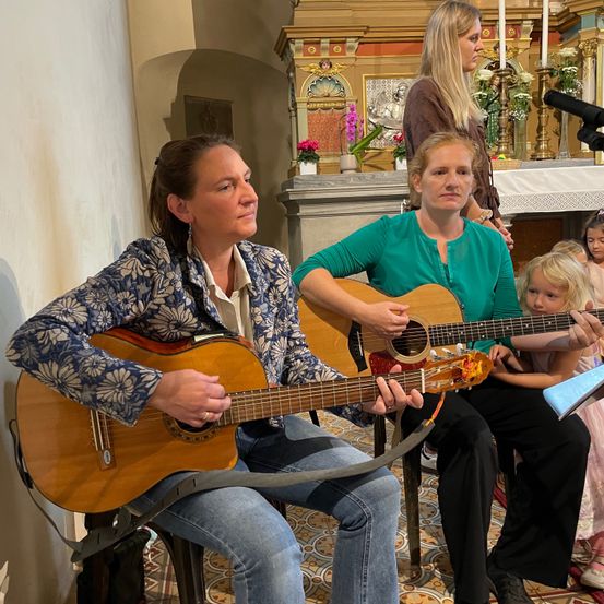 Zwei Frauen spielen Gitarre in einer Kirche. Eine trägt ein Blumenhemd, die andere grün. Ein Kind hält ein Musikblatt vor sich. Dahinter zieren eine Statue und Blumen den Altar.