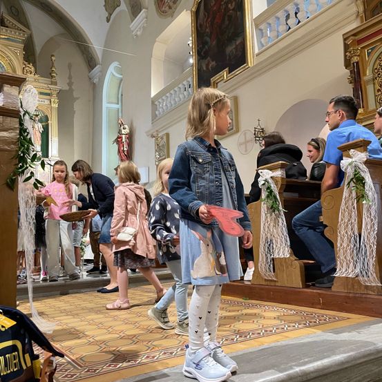 Eine Gruppe von Kindern, einige mit Körben, steht in einem Kirchengang. Ein Kind, das eine Jeansjacke trägt, hält ein rosa Herz. Hinter ihnen sitzen andere auf Kirchenbänken. Eine Statue befindet sich an der Wand.
