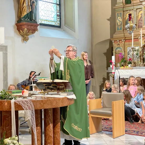 Ein älterer Priester in einer grünen Robe steht an einem Altar und hält eine kleine Hostie. Kinder sitzen auf einem Teppich und Stühlen und beobachten ihn. Eine Frau steht hinter ihm und lächelt. Eine Statue und ein Fenster mit Buntglas befinden sich an der Wand.