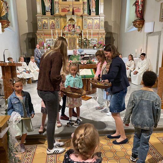 Eine Gruppe von Kindern in einer Kirche hält Blumenkörbe. Zwei Frauen lächeln und halten Körbe. Dahinter sitzen Menschen auf einer Bank und ein Priester steht vor einem Altar.
