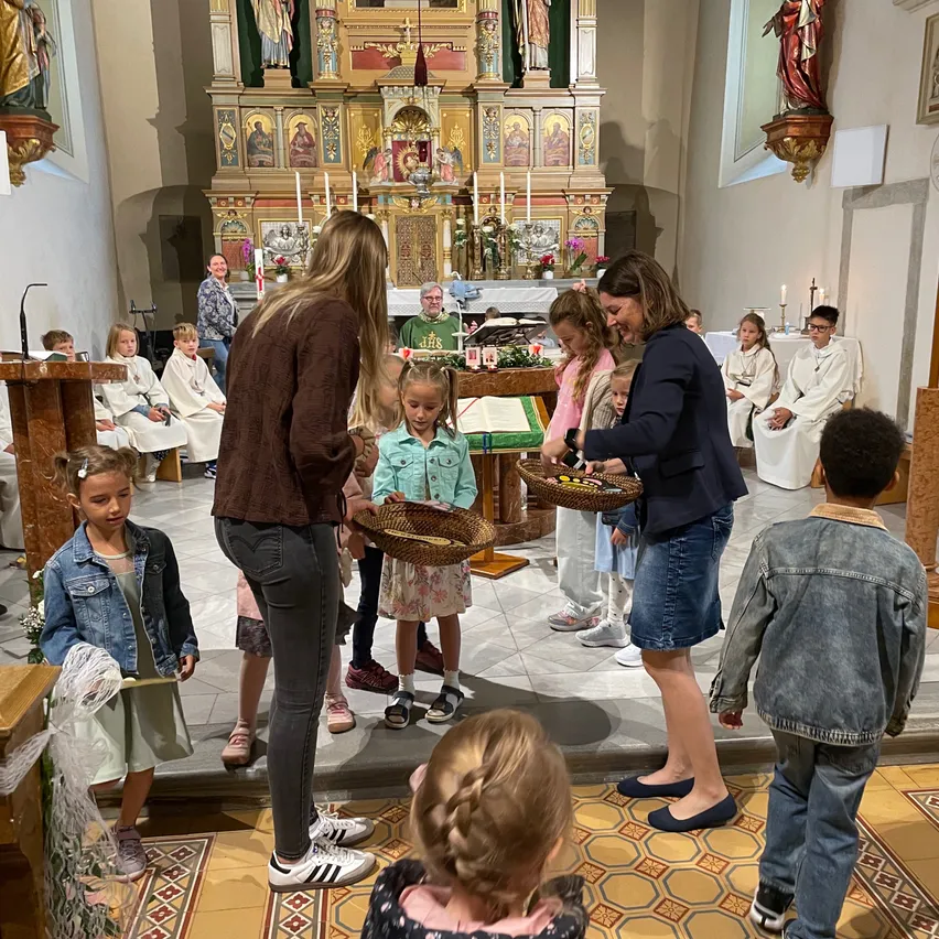 Eine Gruppe von Kindern in einer Kirche hält Blumenkörbe. Zwei Frauen lächeln und halten Körbe. Dahinter sitzen Menschen auf einer Bank und ein Priester steht vor einem Altar.